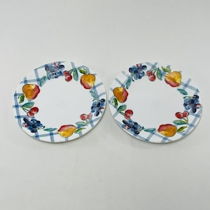 May include: Two round ceramic plates with a white and blue checkered border. Each plate features a painted design of various fruits, including apples, cherries, and grapes, arranged in a circular pattern around the center.