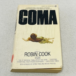 Può includere: Una copertina di un libro tascabile con il titolo "COMA" in grandi lettere nere. La copertina presenta un dipinto di una persona sospesa da delle corde, con il nome dell'autore "ROBIN COOK" e lo slogan del libro "Il thriller medico numero 1 dell'anno..." sotto il titolo.
