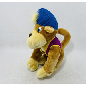 May include: A brown plush monkey wearing a blue hat and a purple vest. The monkey is sitting on its haunches with its tail curled around its body.