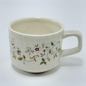 以下が含まれることがあります： 緑、茶色、青の色合いの花柄が施された白いセラミック製のコーヒーカップ。カップは丸みを帯びた形をしていて、持ち手があります。