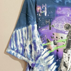 Op de afbeelding: Tie-dye T-shirt met een Star Wars-afbeelding. Het shirt heeft een ruimtegevechtsc&egrave;ne met ruimteschepen, astero&iuml;den en een groot paars ruimtestation. De mouwen hebben een blauw-wit tie-dye patroon.