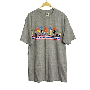 May include: A gray t-shirt featuring a colorful graphic of cartoon characters watching fireworks. The graphic includes red, white, and blue elements, with a row of stars and stripes. The shirt is hanging on a wooden hanger.