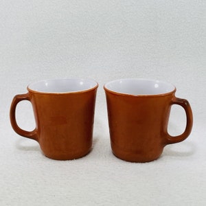 以下が含まれることがあります： 白い内側の茶色のセラミック製のマグカップが2つ。マグカップは丸みを帯びた形をしていて、右側に持ち手があります。