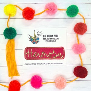 May include: A red felt patch with the word "Hermosa" embroidered in green thread. The patch is surrounded by a white border and has a sew-on and iron-on option. The patch is displayed on a white background with a garland of colorful pom-poms.