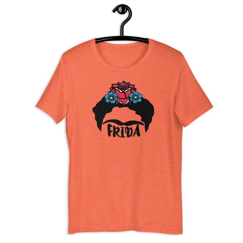 May include: Orange t-shirt with a black and white illustration of Frida Kahlo's face with a red rose and blue flowers in her hair. The text "FRIDA" is printed below the illustration.