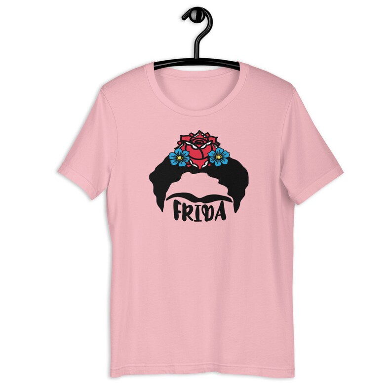 May include: Pink t-shirt with a black and white illustration of Frida Kahlo's face with a red rose and blue flowers in her hair. The text "Frida" is printed below the illustration.