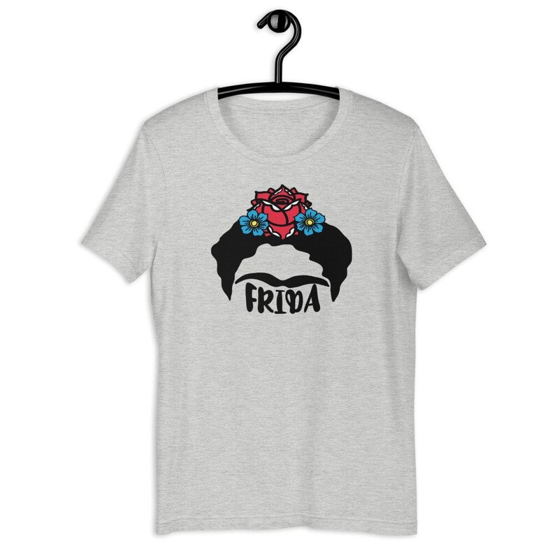 May include: A heather grey t-shirt with a black and white illustration of Frida Kahlo's face with a red rose and blue flowers in her hair. The text "Frida" is printed below the illustration.