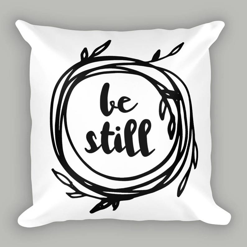 K&ouml;nnte beinhalten: Ein wei&szlig;es Kissen mit schwarzem Text, der "be still" innerhalb eines schwarzen Kranzes lautet.