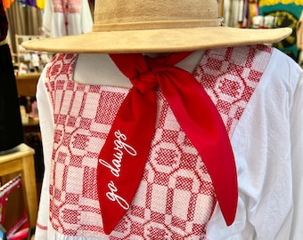 May include: A tan suede hat with a wide brim sits atop a white dress with red embroidered details. A red scarf with the words "go dawgs" in white script is tied around the neck. The dress has a square neckline and a ruffled hem.