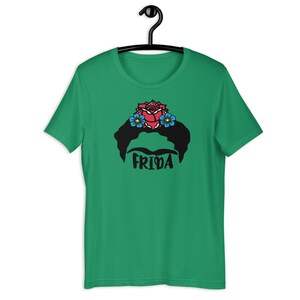 May include: A green t-shirt with a black and white illustration of Frida Kahlo's face with a red rose and blue flowers in her hair. The text "FRIDA" is printed below the illustration.