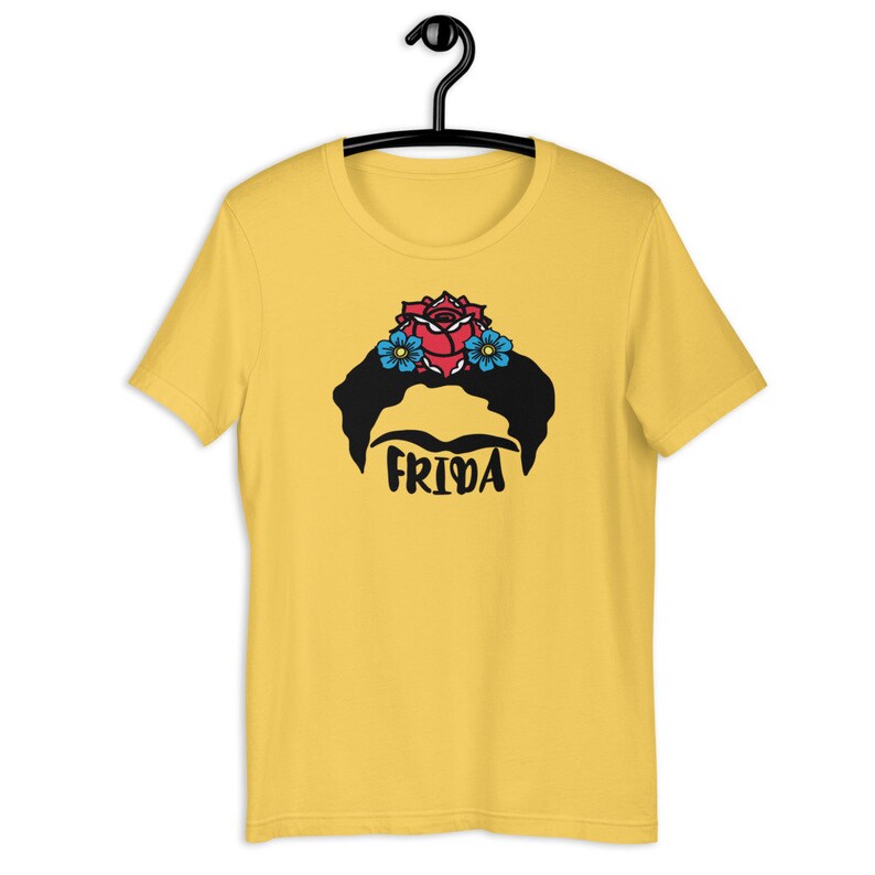 May include: Yellow short-sleeved t-shirt with a black and white illustration of Frida Kahlo's face with a red rose and blue flowers in her hair. The text "Frida" is printed below the illustration.