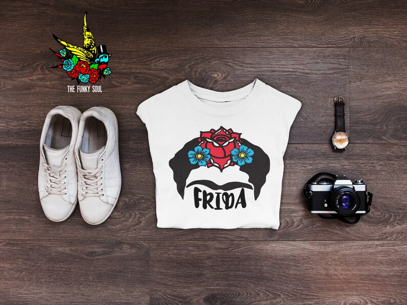 May include: White t-shirt with a black and white illustration of Frida Kahlo with a red rose and blue flowers in her hair. The text "Frida" is printed below the illustration.