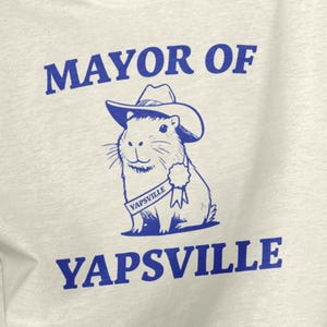 May include: A natural-colored XL t-shirt with the text "MAYOR OF YAPSVILLE" in blue. A cartoon capybara wearing a cowboy hat is in the center. The shirt also has the Legendary Wares logo.