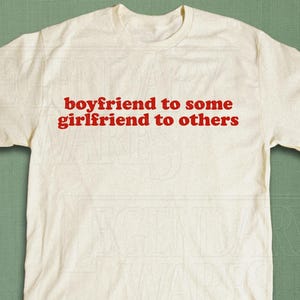 May include: Cream-colored t-shirt with the red text "boyfriend to some girlfriend to others." The shirt is a classic crew neck style. The bottom right corner of the shirt has the text "LEGENDARY WARES."