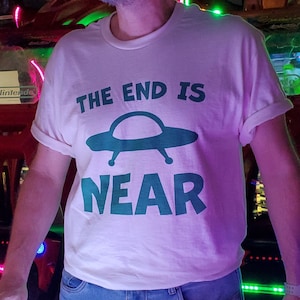 May include: A natural-colored t-shirt with the text "THE END IS NEAR" and a teal UFO graphic. The shirt is size large. The person in the image is 5'10" tall.