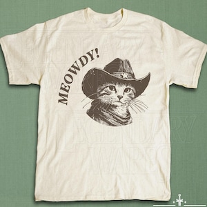 May include: Cream-colored t-shirt featuring a sepia-toned illustration of a cat wearing a cowboy hat and a neckerchief. The text "MEOWDY!" is printed above the cat's head. The shirt has short sleeves and a crew neck.