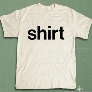 May include: Cream-colored t-shirt with the word "shirt" printed in bold black letters across the chest. The shirt is made of a soft material and has short sleeves. The Legendary Wares logo is at the bottom.