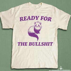 以下が含まれることがあります： 紫色の「READY FOR THE BULLSHIT」の文字と紫色のフェレットのイラストがプリントされた、クリーム色のTシャツ。カジュアルな普段着としてデザインされています。