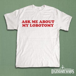 May include: White t-shirt with the red text "ASK ME ABOUT MY LOBOTOMY". The shirt is a crew neck style and appears to be made of cotton. The bottom right corner of the shirt has the text "LEGENDARY WARES" in a stylized font.