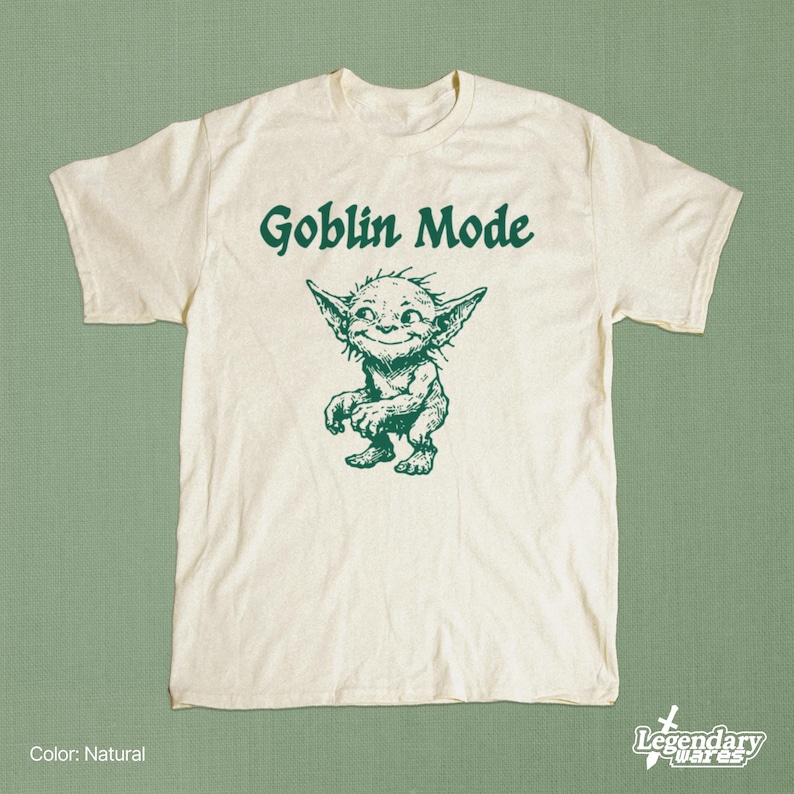 May include: Natural-colored t-shirt with the words "Goblin Mode" above a green illustration of a goblin. The goblin is smiling and has pointed ears. The shirt is from Legendary Wares.