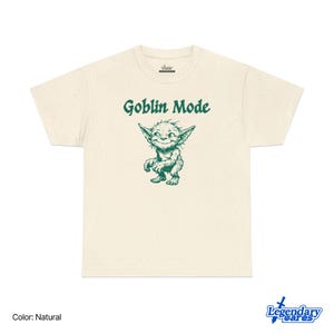 May include: Natural-colored t-shirt with the text "Goblin Mode" above a green cartoon goblin illustration. The goblin has large pointed ears and a mischievous grin. The shirt is made of a soft material.