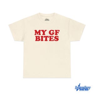 May include: Cream-colored t-shirt with the text "MY GF BITES" in bold red letters. The shirt has a classic crew neck and short sleeves. The shirt is made of a soft, speckled fabric.