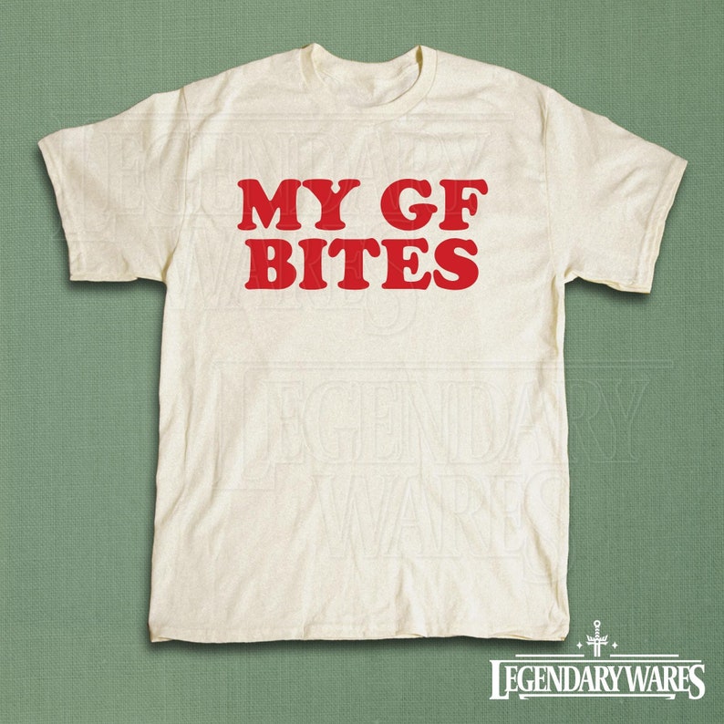 May include: Cream-colored t-shirt with the red text "MY GF BITES" in a bold, sans-serif font. The shirt has short sleeves and a crew neck. The Legendary Wares logo is visible at the bottom.