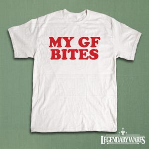 May include: White t-shirt with the red text "MY GF BITES". The shirt is made of a soft material and has short sleeves. The shirt is displayed on a green background.
