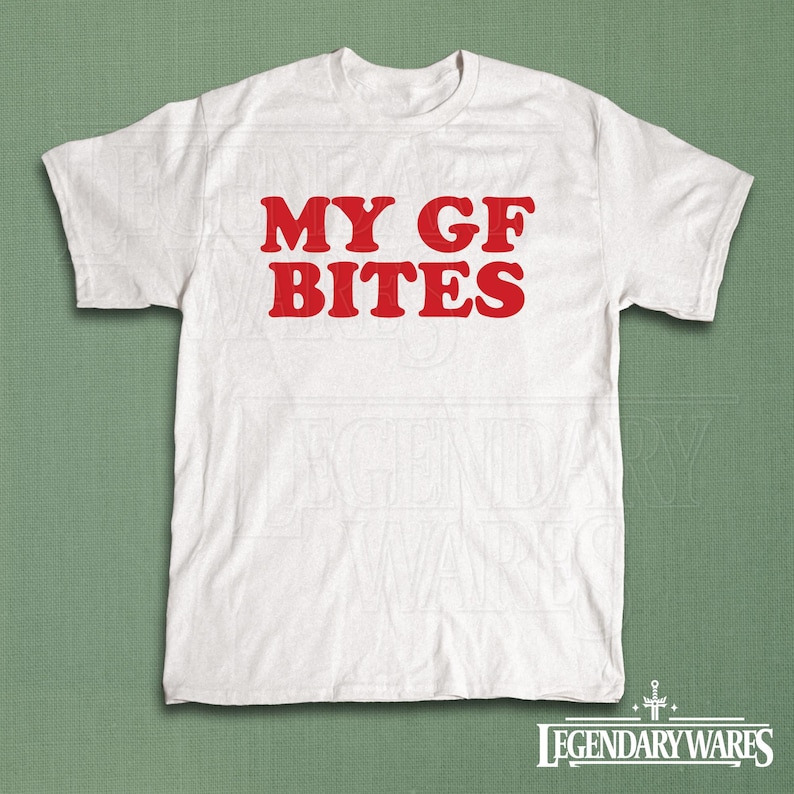 May include: White t-shirt with the red text "MY GF BITES". The shirt is made of a soft material and has short sleeves. The shirt is displayed on a green background.