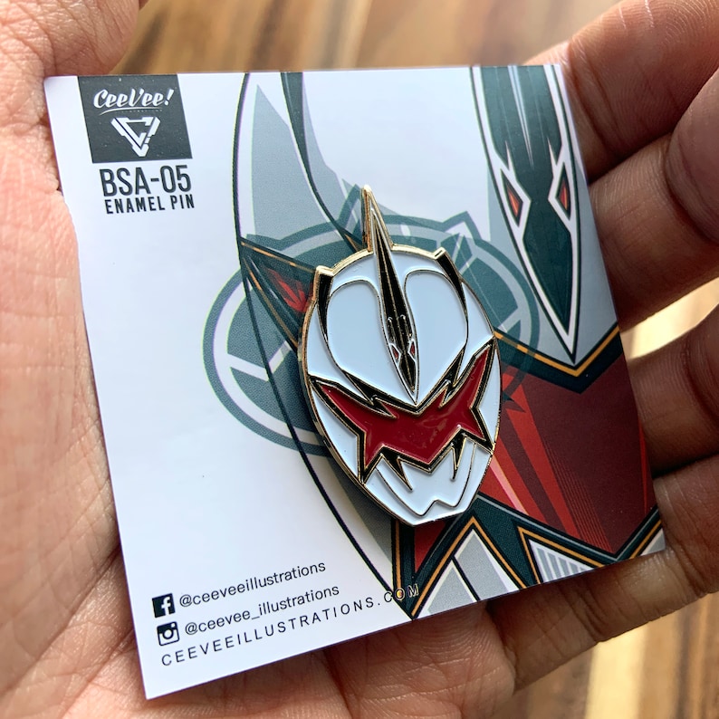 May include: A white and red enamel pin with a gold outline. The pin features a stylized mask with a red and black design. The pin is labeled "BSA-05 Enamel Pin" and "CeeVee!" with a logo. The pin is on a white card with social media information.
