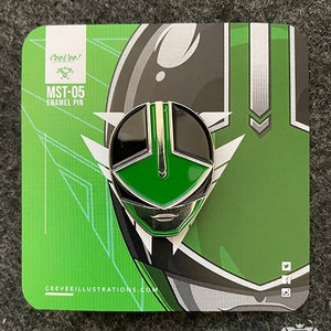 May include: Green and black enamel pin featuring a stylized face with a green visor and a black background. The pin is on a card with the text "Cooltee! MST-05 Enamel Pin" and the website "ceeveeillustrations.com".