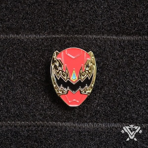 May include: A red and gold enamel pin featuring a stylized face with a black mouth and a blue diamond in the center. The pin is a collectible for fans of the Power Rangers franchise.