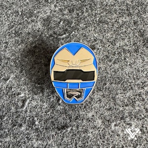 Puede incluir: Un pin metálico azul y plateado con un diseño de casco estilizado. El pin tiene un borde azul con detalles plateados y negros, y un emblema plateado en la parte inferior. El pin está sobre un fondo gris texturizado.