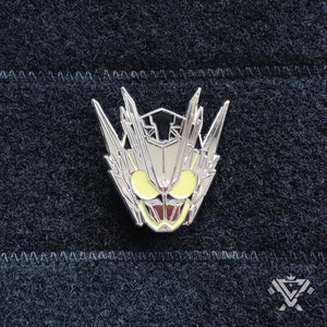 May include: A silver-colored metal pin featuring a stylized face with sharp, angular details. The pin has yellow eyes and a red mouth, set against a dark background. The design is intricate and detailed.