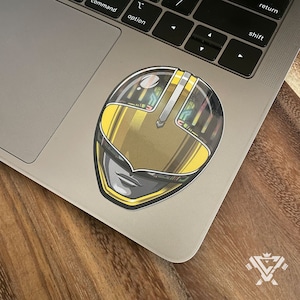 May include: A yellow and black Power Rangers helmet sticker with a detailed design. The helmet has a visor and a face with a serious expression.