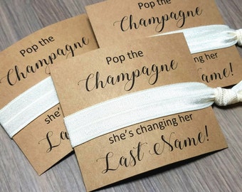 Puede incluir: Tres tarjetas marrones con gomas elásticas blancas para el pelo. Las tarjetas tienen el texto "Pop the Champagne" y "she's changing her last name!" impreso en ellas.