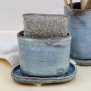 Puede incluir: Dos recipientes de cerámica hechos a mano para utensilios de cocina en tonos azules y marrones. Uno contiene un estropajo de acero inoxidable, el otro utensilios de madera. Los recipientes están sobre platillos a juego. Una toalla blanca en el fondo.