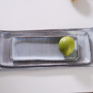 May include: Three rectangular ceramic trays nested together, featuring a light grey glaze with a vertical ribbed texture and dark brown edges. A single bright green lime rests on the surface of the smallest tray, highlighting the scale of the set.