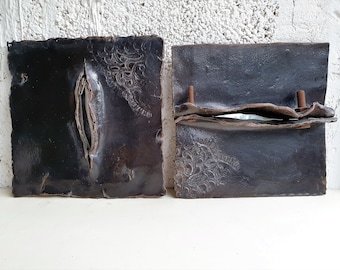 May include: Two dark, rectangular ceramic pieces with a distressed finish. One features a zipper detail, the other has a raised, decorative floral pattern and is held together with bolts. The pieces are likely part of a larger art installation.