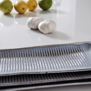 May include: A stack of rectangular ceramic serving platters with a textured, ridged surface in shades of grey and blue. Limes, lemons, and garlic cloves are scattered on the white surface in the background. The platters are stacked on top of each other.