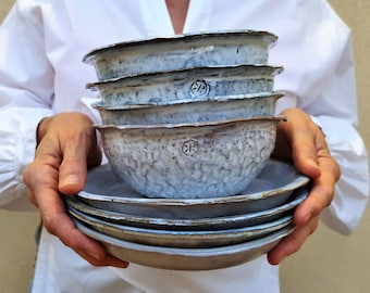 May include: A stack of handmade ceramic bowls and plates. The bowls and plates are white with a textured surface and a brown rim. The bowls have a small, embossed design. The person holding the bowls is wearing a white shirt.