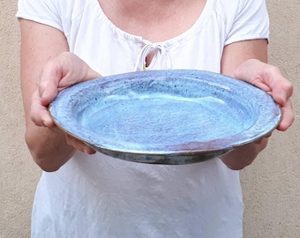 May include: A handmade ceramic plate with a wide rim. The plate features a mottled blue glaze with hints of purple and brown. The plate is round and appears to be approximately 12 inches in diameter, suitable for serving food.