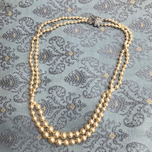 Puede incluir: Un collar de perlas de dos hilos con un cierre decorativo plateado. Las perlas son de color blanco roto cremoso y varían en tamaño, creando un efecto en capas. El collar descansa sobre una tela azul estampada.