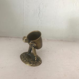 May include: A small brass figurine of a person carrying a bucket on their back. The figurine is a decorative trinket or a small container.