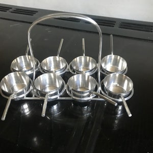 Op de afbeelding: Een zilveren metalen dienblad met acht kleine kommen en een handvat. De kommen zijn gerangschikt in twee rijen van vier. Het dienblad is ontworpen om kleine porties voedsel te serveren.