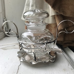 May include: A silver and glass butter dish with a clear glass jar and a lid. The dish has a silver base with a scalloped edge and a silver handle on each side. The dish also has a silver spoon with a decorative handle.