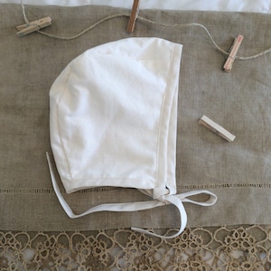 May include: A white cotton bonnet with a simple design and tied strings. The bonnet is lying on a brown linen fabric with lace trim.