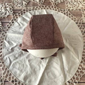 May include: A brown linen bonnet with a simple design. The bonnet is on a white fabric background.