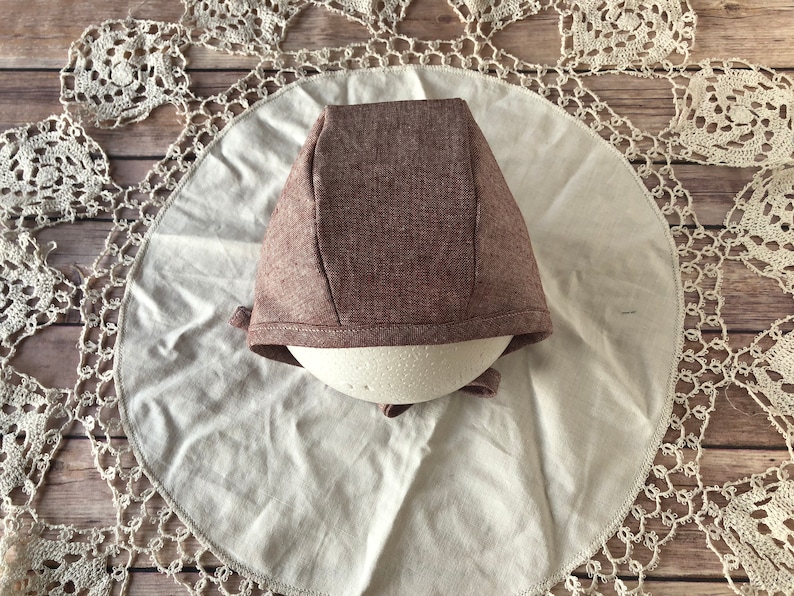 May include: A brown linen bonnet with a simple design. The bonnet is on a white fabric background.