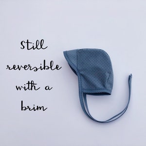 May include: A blue baby bonnet with white polka dots and ties. The bonnet is reversible and has a brim. The text "still reversible with a brim" is written on a white background.